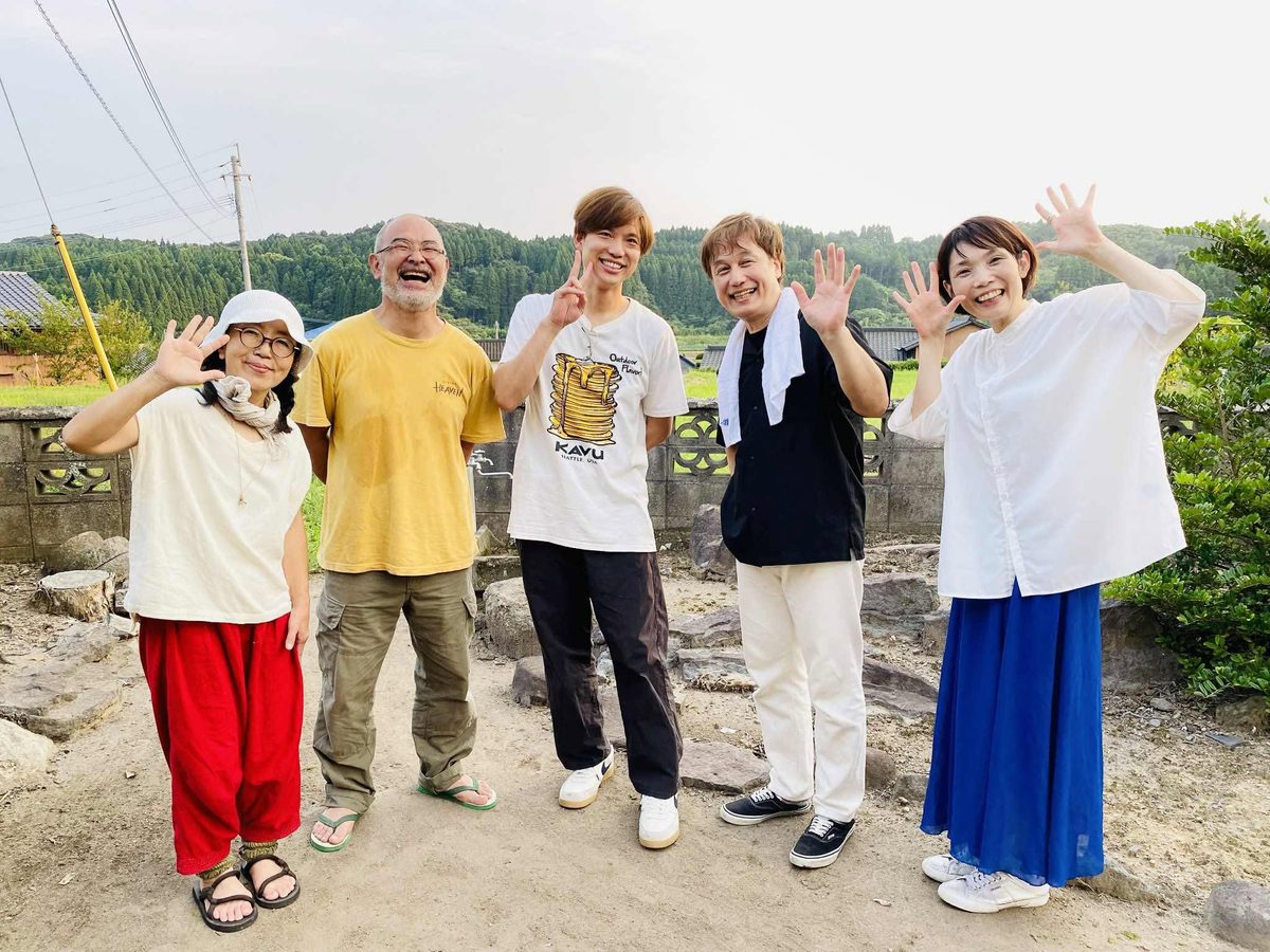 緑の山と田んぼを背に、五人が手を振って笑っている