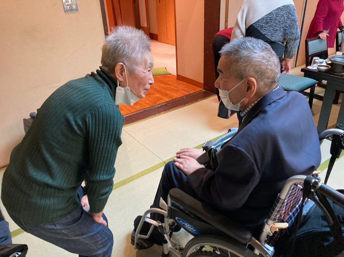 旅館の座敷で、旧友が腰を深くかがめ、車いすの男性と目線を合わせて話し込んでいる