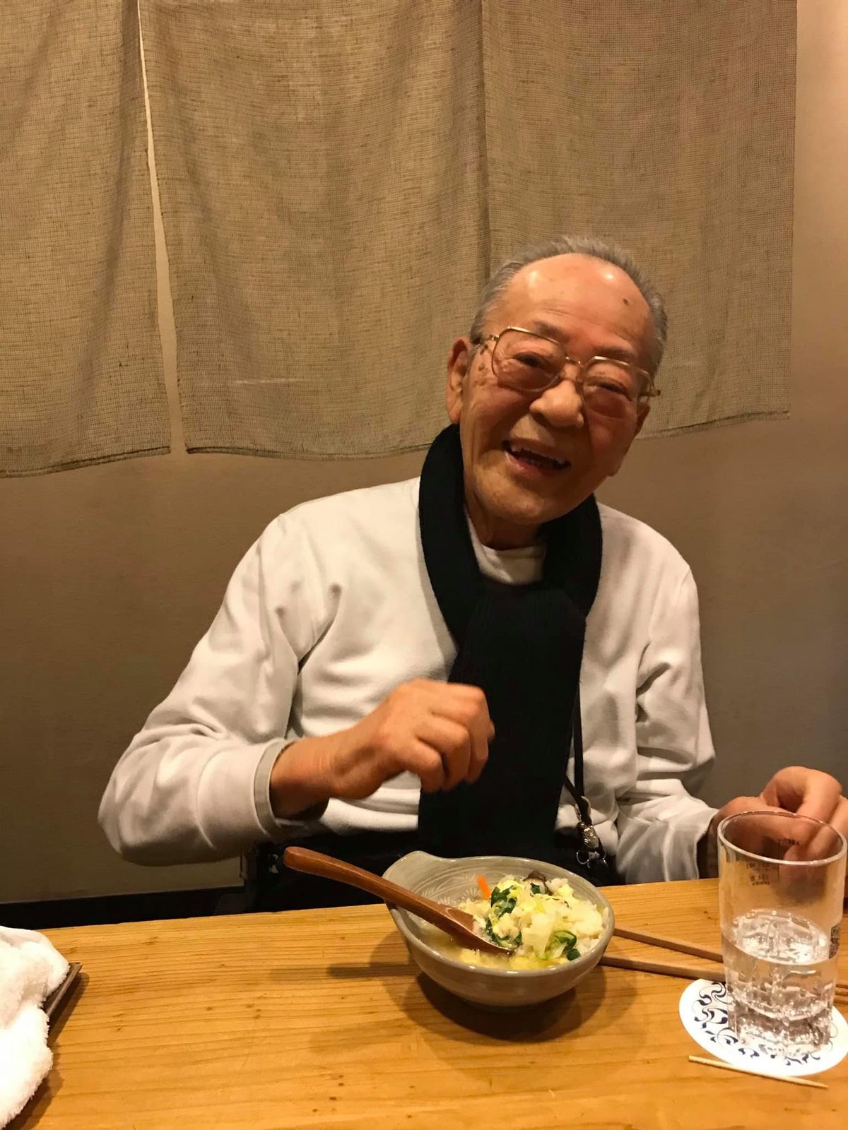 店ののれんの前の席で、拳を握って満面の笑顔を見せる高齢の男性